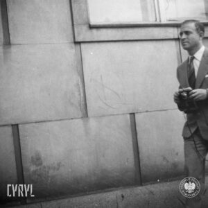 mężczyzna w garniturze z aparatem fotograficznym stoi na tle ściany budynku