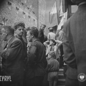 grupa ludzi, przeważnie młodych mężczyzn i dzieci na schodach prowadzących do budynku, o ścianę budynku oparte flagi, w głębi fragment okazałego gmachu