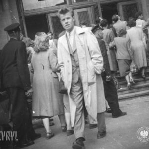 grupa kobiet i mężczyzn wchodzi po schodach do budynku, w głębi otwarte drzwi do budynku, na pierwszym planie młody mężczyzna w jasnym prochowcu wychodzi z budynku