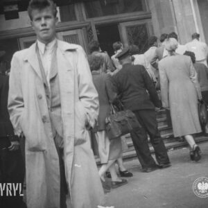grupa kobiet i mężczyzn wchodzi po schodach do budynku, w głębi otwarte drzwi do budynku, na pierwszym planie młody mężczyzna w jasnym prochowcu stoi przed budynkiem