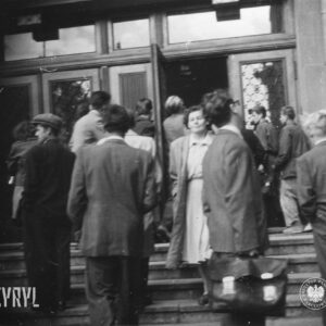 grupa kobiet, mężczyzn i kilku chłopców wchodzi po schodach do budynku, w głębi otwarte drzwi do budynku