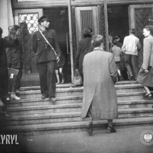 grupa kobiet, mężczyzn i kilku chłopców wchodzi przez otwarte drzwi do budynku, na schodach stoi mężczyzna w mundurze (prawdopodobnie MPK) z torbą przewieszoną przez ramię