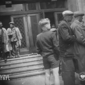 dwóch mężczyzn wychodzi z budynku, z prawej kilku mężczyzn, wśród nich chłopiec