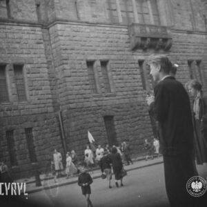 kilka osób przed okazałym gmachem, część przechodzi przez ulicę, po drugiej stronie , na chodniku stoi kilkanaście osób, dwie flagi oparte o elewację budynku