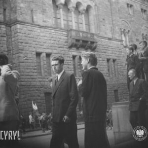 na pierwszym planie stoi kilka osób, w głębi kilka osób zgromadzona przed okazałym gmachem, dwóje dzieci trzyma flagi