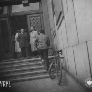 dwóch mężczyzn stoi przed drzwiami do budynku, na pierwszym planie mężczyzna wchodzi po schodach, o ścianę budynku oparty rower