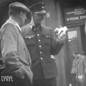 dwóch mężczyzn, mężczyzna po prawej w mundurze, oglądają elementach urządzenia, za nimi wejście do budynku, nad wejściem tablice informacyjne