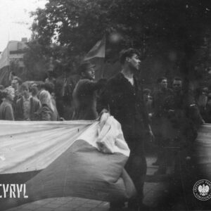 kilku młodych mężczyzn niesie dużą flagę, otaczają ich ludzie zgromadzenie na ulicy, w głębi budynek mieszkalny