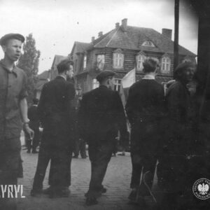 młodzi mężczyźni stoją na ulicy, w głębi budynek mieszkalny z mansardowym dachem