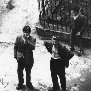 dwóch młodych mężczyzn i chłopiec sfotografowani od góry, prawdopodobnie z okna budynku,  stoją przy metalowym ogrodzeniu