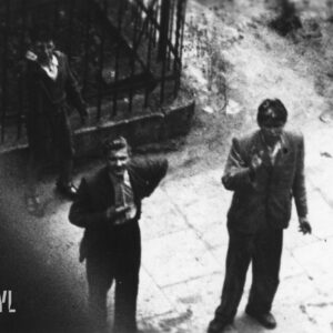 dwóch młodych mężczyzn i chłopiec sfotografowani od góry, prawdopodobnie z okna budynku,  stoją przy metalowym ogrodzeniu