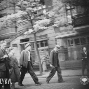 kilku mężczyzn idzie całą szerokością brukowanej ulicy, wzdłuż ulicy kamienice