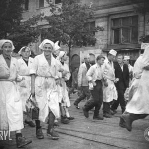 na pierwszym planie grupa kobiet w białych kitlach i chustkach na głowach, część z nich trzyma w rękach torby, w głębi kilku mężczyzn i chłopców, część w białych kitlach i fartuchach oraz białych czepkach na głowie, w głębi kamienice