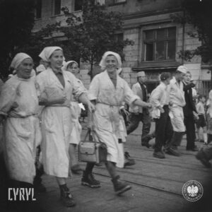 na pierwszym planie grupa kobiet w białych kitlach i chustkach na głowach, część z nich trzyma w rękach torby, w głębi kilku mężczyzn i chłopców, część w białych kitlach i fartuchach oraz białych czepkach na głowie, w głębi kamienice