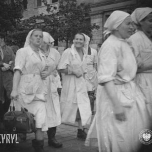 na pierwszym planie grupa kobiet w białych kitlach i chustkach na głowach, jedna z nich trzyma w ręku torbę, za nimi kilku mężczyzn w roboczych ubraniach