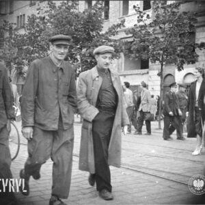 grupy ludzi, przeważnie mężczyzn, część w roboczych ubraniach idzie ulicą, w głębi niewielkie drzewa, za nimi kamienice 