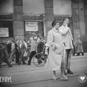 na pierwszym planie kobieta i mężczyzna stoją na ulicy, w głębi grupa ludzi idzie chodnikiem, częściowo ulicą, za nimi budynek