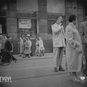 na pierwszym planie kobieta i mężczyzna stoją na ulicy, w głębi kilka osób idzie chodnikiem, za nimi budynek