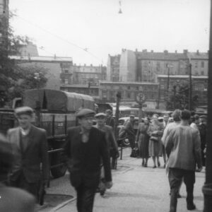 grupy ludzi i pojedynczych osób, część stoi, część idzie, przy ulicy, z lewej stoi samochód ciężarowy, w głębi kilkupiętrowe kamienice