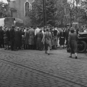 brukowana ulica, przy niej zgromadzenie ludzi, w głębi fragment budynku kościoła