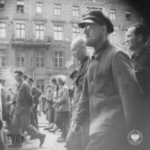 duża grupa kobiet i mężczyzn idzie ulicą, w głębi kamienica, z kilku okien wyglądają ludzie