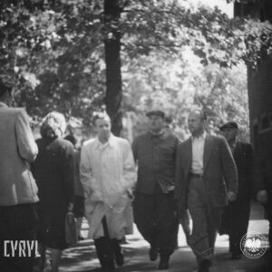 grupa ludzi idzie chodnikiem, z prawej fragment budynku, w głębi drzewa