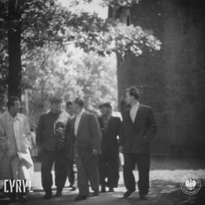 grupa ludzi idzie chodnikiem, z prawej fragment budynku, w głębi drzewa