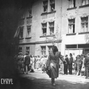 duża grupa ludzi stoi na chodniku, na pierwszym planie kobieta w chustce na głowie przechodzi przez ulicę, w głębi kamienice, w niektórych oknach stoją ludzie i obserwują ulicę