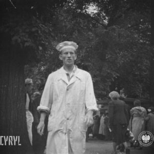 na pierwszym planie mężczyzna w białym kitlu i białym czepku na głowie, w głębi grupa ludzi stoi przy kiosku handlowym, wysokie drzewa