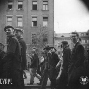 duża grupa, przeważnie mężczyzn, idzie ulicą, w głębi kamienice, w kilku oknach ludzie obserwują ulicę