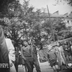 grupy ludzi na placu, w głębi drewniane ogrodzenia i stoiska oraz drzewa, zza drzew widoczne kilkupiętrowe budynki