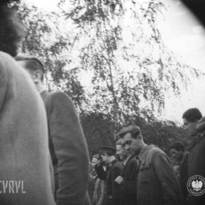 grupa ludzi, ujętych od pasa w górę idzie ulicą, w głębi krzaki i niewielkie drzewa