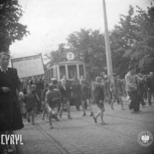 duża grupa ludzi idzie brukowaną ulicą, w głębi tramwaj, po obu stronach ulicy drzewa