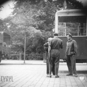trzech mężczyzn, dwóch w roboczych kombinezonach stoją na środku brukowanej ulicy, z prawej fragment wagonu tramwajowego, w głębi drzewa