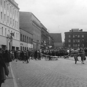 tłum ludzi na całej szerokości ulicy, w głębi, na podwyższeniu kilkanaście osób z flagami, po obu stronach ulicy i na wprost kilkupiętrowe budynki
