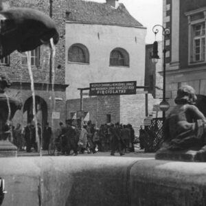grupa ludzi na narożniku ulic, po obu stronach kamienice, na pierwszym planie fragment fontanny