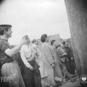 grupa ludzi, wśród nich kobieta w sukni w kratkę i z siatką w dłoni, na pierwszym planie mężczyzna w roboczym ubraniu, poplamionym farbą