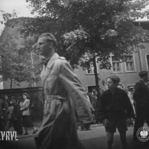 grupy ludzi idą lub stoją na ulicy, w głębi trzykondygnacyjny niewielki budynek mieszkalny, przed nim pośrodku drzewo, na pierwszym planie idzie mężczyzna w jasnym płaszczu