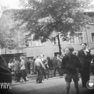 grupy ludzi stoją na ulicy, w głębi trzykondygnacyjny, niewielki budynek mieszkalny, przed nim pośrodku drzewo