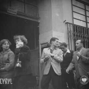 grupa ludzi idzie wzdłuż budynku, po lewej otwarta brama