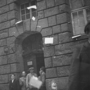 kilku mężczyzn stoi w drzwiach wejściowych do budynku, z okien lecą kartki papieru