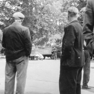 dwóch mężczyzn stoi tyłem, trzymając ręce w kieszeniach, w głębi samochody wojskowe, za nimi drzewa