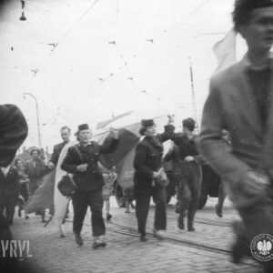 dwie kobiet w mundurach i furażerkach na głowach niesie flagę, idąc brukowaną ulicą, wokół nich duże grupy ludzi