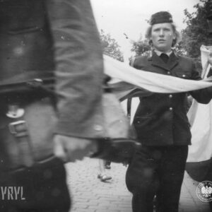 kobieta w mundurze i furażerce na głowie trzyma w rękach flagę, idąc brukowaną ulicą, wokół niej duże grupy ludzi