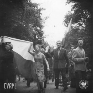 dwie kobiety w mundurach oraz w furażerkach na głowie, młoda dziewczyna w sukience oraz mężczyzna niosą duża flagę, idąc brukowaną ulicą, obok nich idzie kilka osób