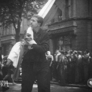 na pierwszym planie mężczyzna w roboczym ubraniu z flagą na ramieniu, w głębi grupa ludzi na tle ozdobnej willi miejskiej
