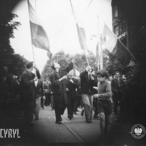 duża grupa ludzi idzie brukowaną ulicą, na przodzie kilku młodych mężczyzn niesie polskie flagi na drzewcach, wyraźna czarna obwódka wokół zdjęcia