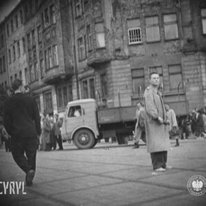 skrzyżowanie ulic, na chodniku grupy ludzi, w centrum samochód ciężarowy, na pierwszym planie mężczyzna w prochowcu, w głębi narożna kamienica