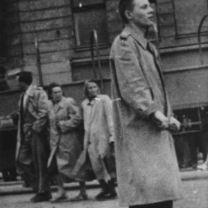 skrzyżowanie ulic, na pierwszym planie mężczyzna w prochowcu , w głębi fragment kamienicy, przed nią chodnikiem idą trzy osoby