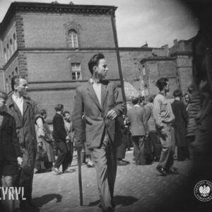 grupa ludzi stoi na skrzyżowaniu ulic, w głębi masywny budynek